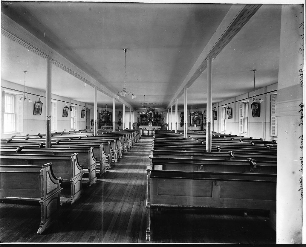 Prise depuis le fond de la salle, cette photographie montre la chapelle de l'asile de Longue Pointe en 1911. La longue salle était remplie de rangées de bancs qui s'étendaient jusqu'au chœur. Des fenêtres, situées de part et d'autre de la salle, alternaient avec des tableaux religieux.