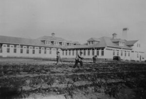 Ancienne photo en noir et blanc représentant des hommes travaillant dans un champ devant un bâtiment institutionnel d'un étage