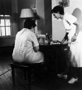 Ancienne photo en noir et blanc d’une femme assise devant une machine à coudre et une autre femme debout à sa droite qui l’assiste