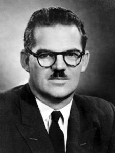 1950s black and white photot of man with dark hair, mustache, glasses where a jacket and a tie.