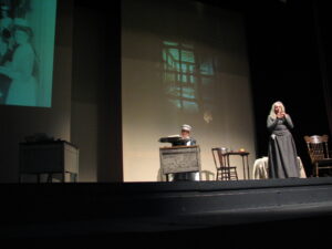 stage performance with woman with long white hair and grey dress standing to stage right and woman with nurse's cap sitting at desk with typewriter