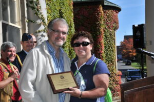 Un homme et une femme tiennent une plaque honorifique devant la façade d'un immeuble