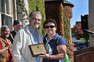man and woman standing smiling in front of buildng holding award plaque