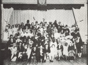 black and white group photograph of approx 100 adults dressed in costumes