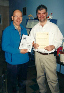 two smiling men standing next to each other holding publications