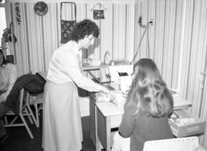 Photo en noir et blanc datant environ des années 1970 avec une femme qui aide une autre femme à utiliser une machine à coudre