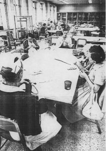 Ancienne photo en noir et blanc représentant des femmes et des hommes dans un atelier institutionel en train de tricoter