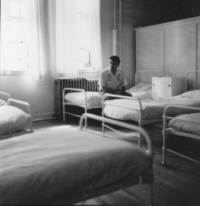 Ancienne photo en noir et blanc représentant un homme assis sur un lit dans un dortoir avec de grandes fenêtres