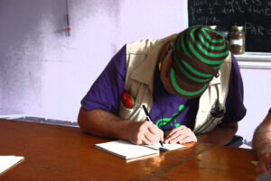 colour photo of person in green cap sitting at table intently writing