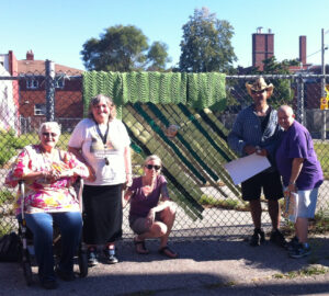 colour photo of smiling group of people standing outside against fence with knitted blanket hung on it