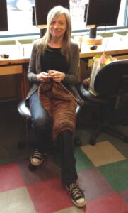 colour photo of smiling young woman with long blond hair sitting in office chair knitting