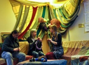 colour photo of people on couch knitting, furniture and skylight are draped with beautiful coloured fabric