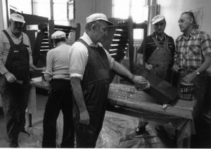 Photo en noir et blanc d'un groupe d'hommes en train de vernir des chaises dans un atelier