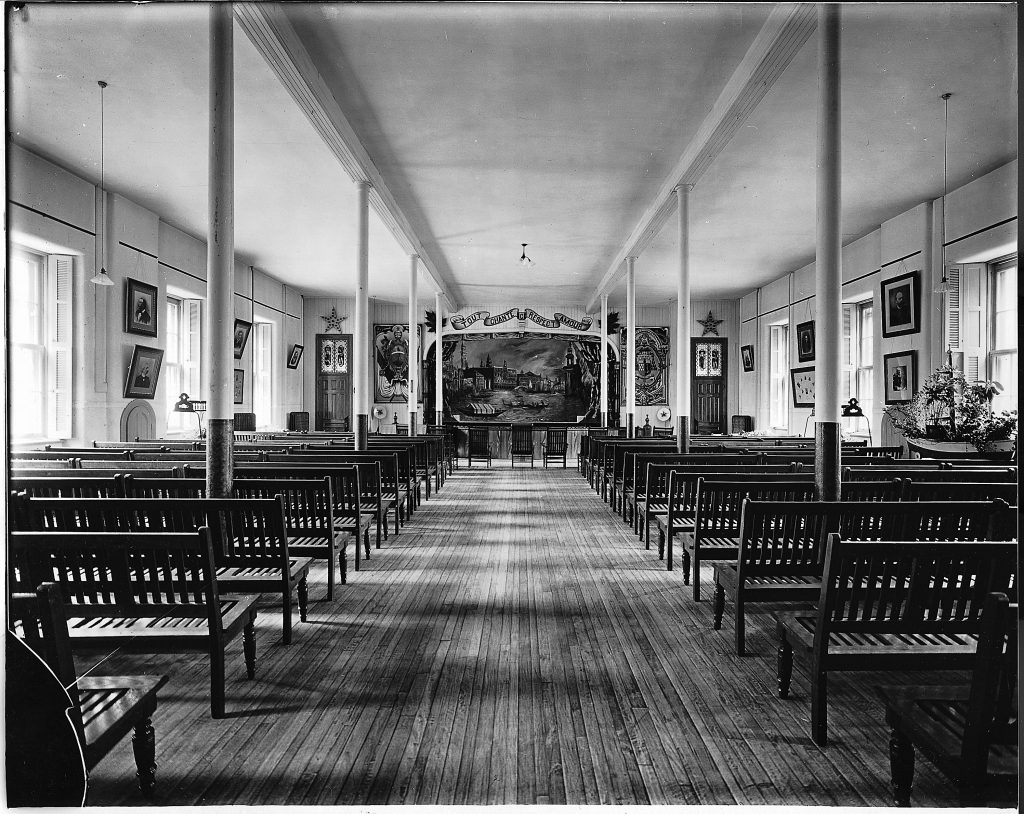 Vus de dos, des bancs en bois bordent la longue salle jusqu’à la scène du théâtre. Des fenêtres sont disposées de part et d’autre de la salle, entrecoupées de portraits. 