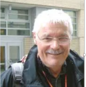 Head and shoulders shot of apparent man with white hair, mustache and glasses smiling.