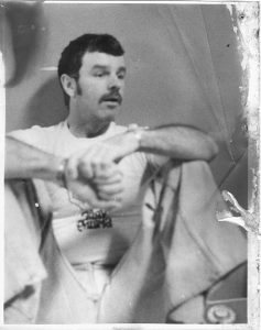 man with dark hair and mustache sitting with his knees up