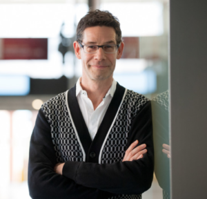 Dark haired apparent man with glasses smiling with arms folded.