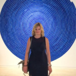 Body shot of blond serious looking apparent woman standing against blue circular wall art.