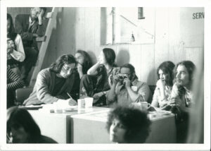 group of 1970s peole sitting around a table in a basement.  One has camera and another is writing something. Others laugh or talk.