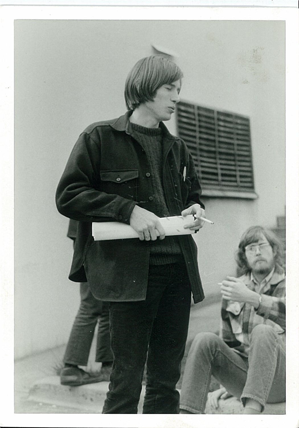 1970s person holding rolled up paper in hand. Another 1970s person sitting on the ground.