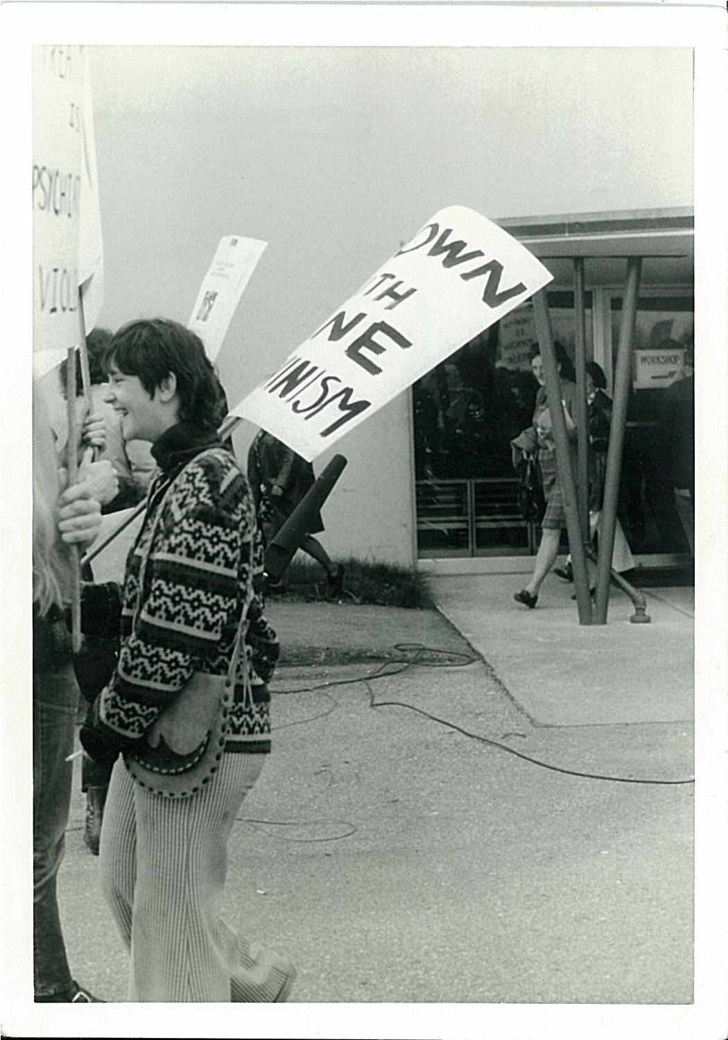 person wearing oversize patterned sweater and carrying protest sign.