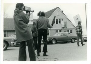 1970s media people with camera and mic, in background one protester with sign, 1970s cars and institutional building