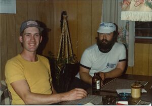 Two people at table: one on left has a yellow tshirt and baseball cap and one on right has white tshirt and cap