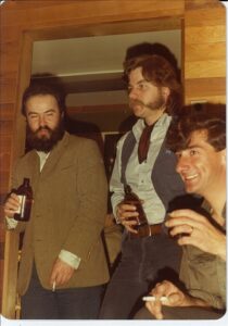 3 people standing and drinking beers: dark haired bearded person on left wearing brown sports jacket, dark haired person in middle with mutton chops, shirt and vest, person on right with dark hair and clean-shaven 