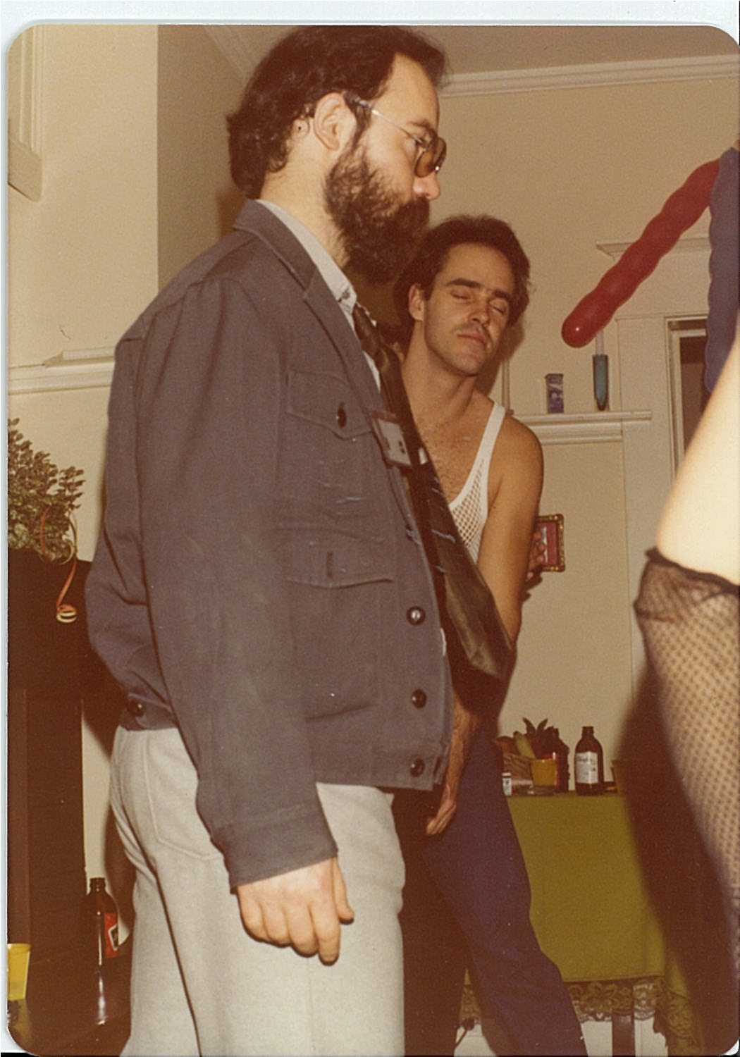 person with dark hair and beard wearing grey jacket, another person with dark hair wearing a tank top behind