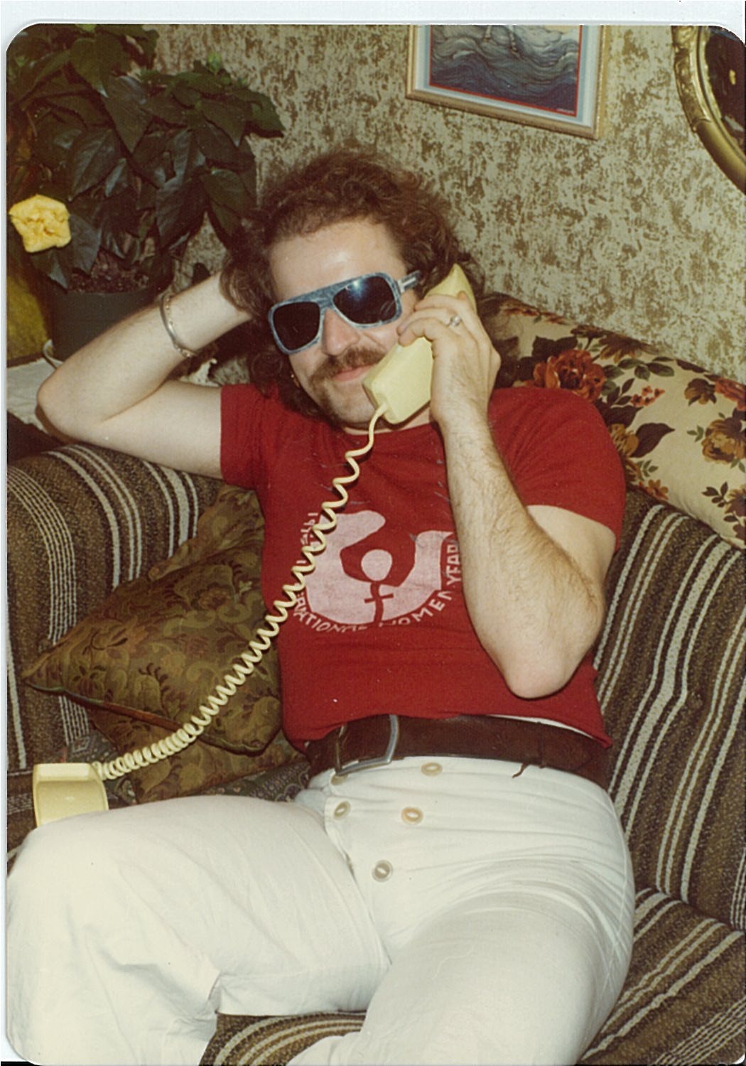 person with dark hair, mustache, white trousers and red tshirt sitting on couch and talkin on phone