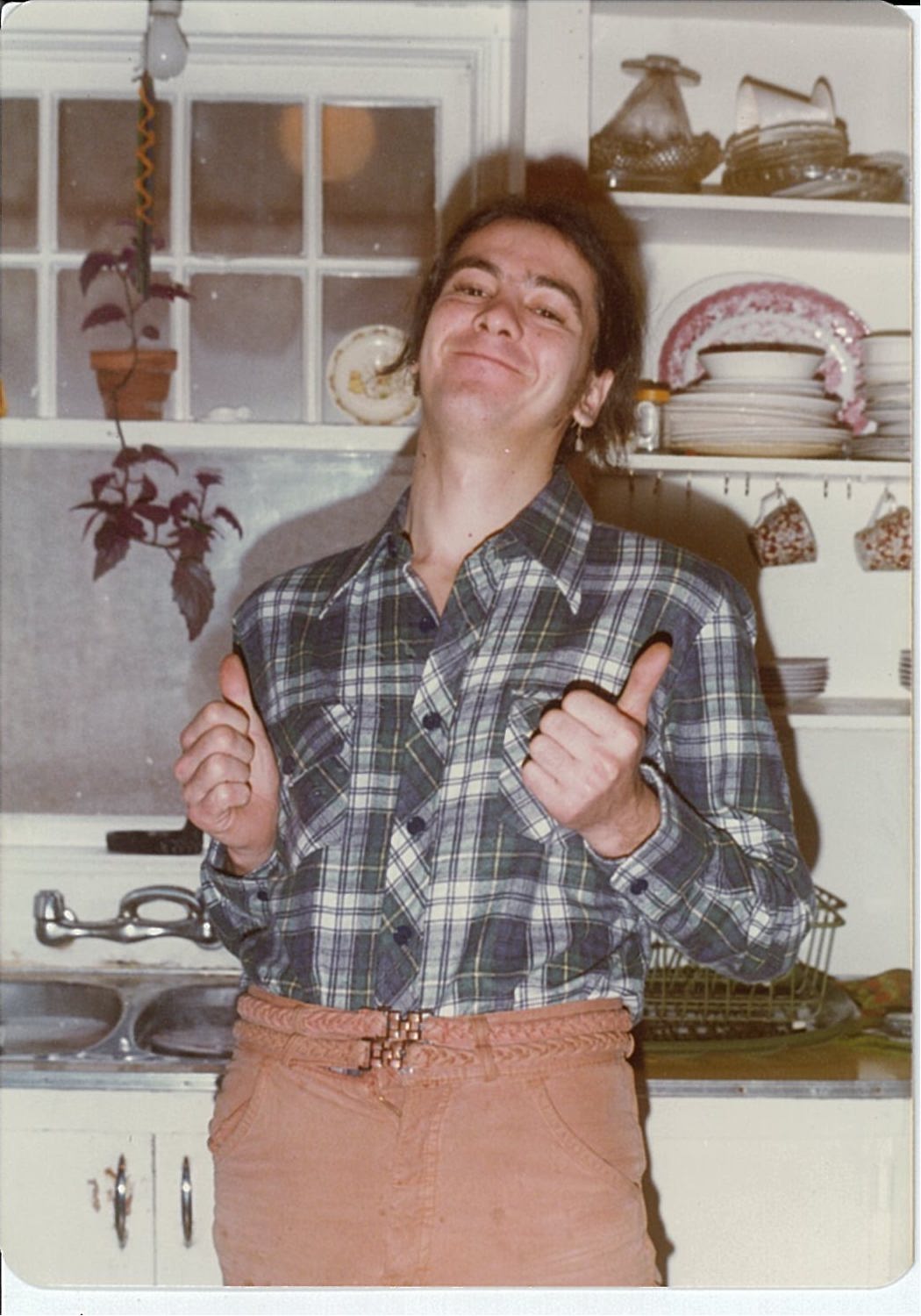 Person with dark hair, plaid shirt and brown trousers standing in front of kitchen sink giving a thumbs up