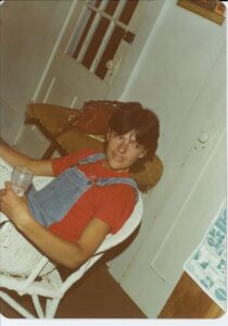 person with short dark hair wearing red tshirt, dunagrees, sitting in a chair