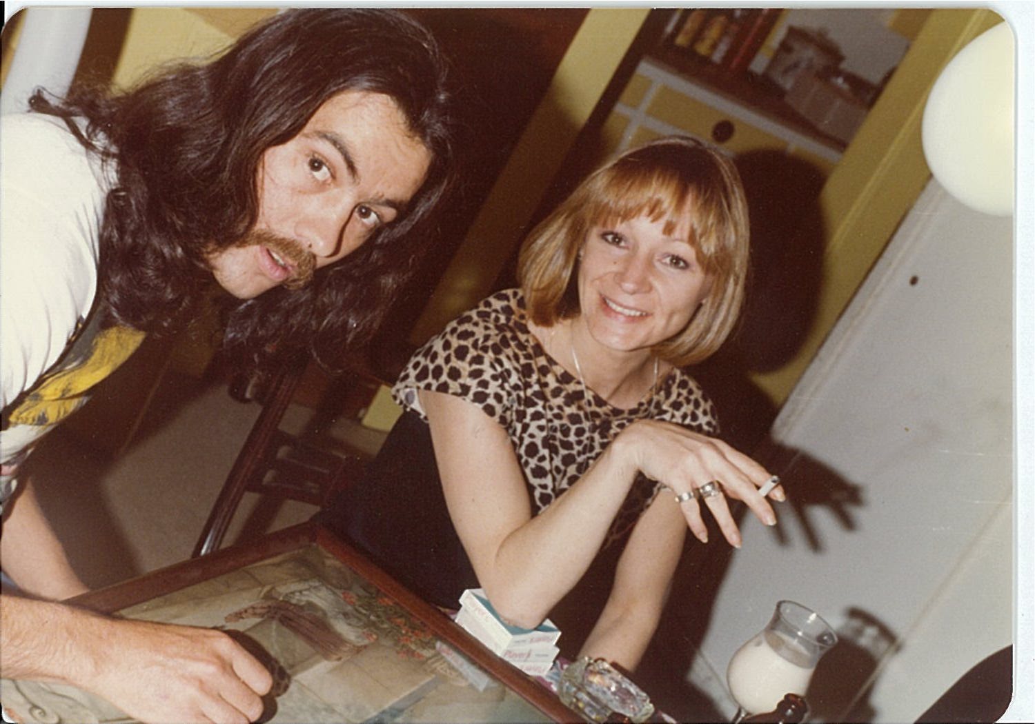 person with long dark hair and mustache leaning over table, person with strawberry blond hair wearing blouse sitting at table