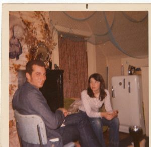 dark haired man and dark hair women sitting in house, fridge behind